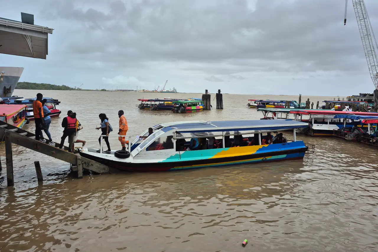Passengers stepping from a wooden landing onto a painted water taxi on the Demerara River in Georgetown, with more boats and a crane behind