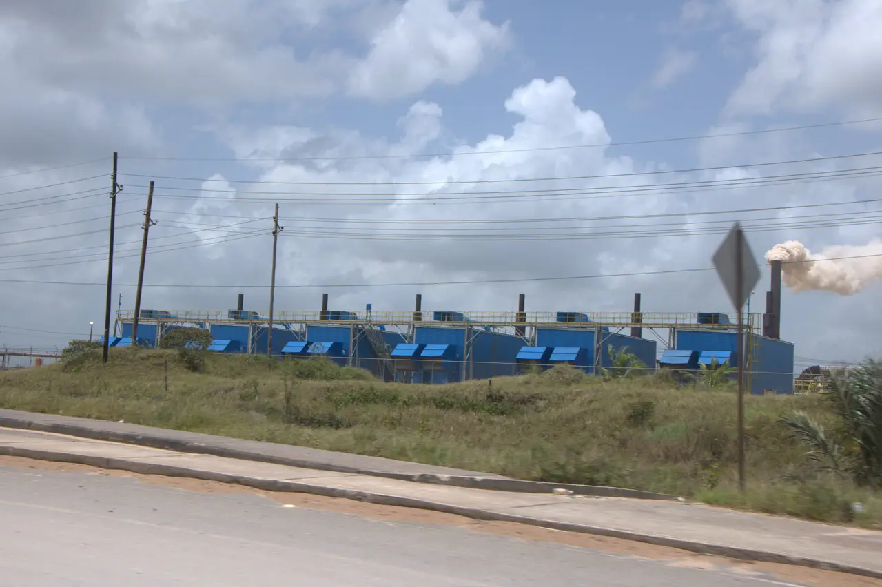 A row of blue containerised generator units behind a fence, with overhead power lines along the road and an exhaust plume