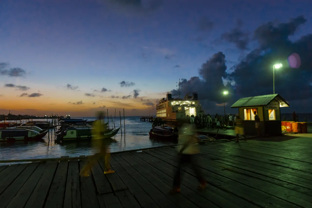 A timber wharf at dusk with people walking, small boats moored alongside and a lit ferry beyond